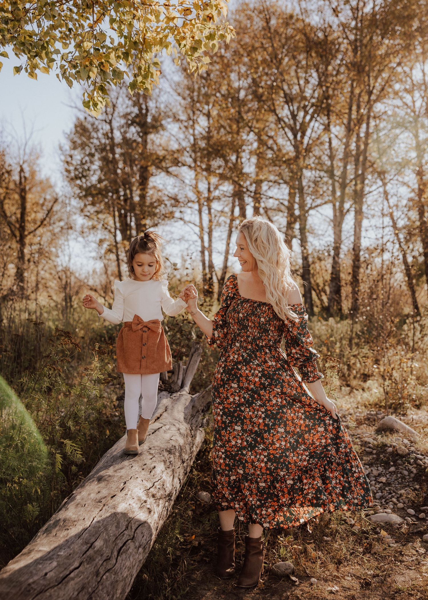 A woman in a floral dress holds a toddler girl's hand as they walk on a fallen log in an autumn forest.