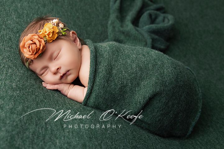 Sleeping newborn wrapped in green blanket wearing orange floral headband on dark green backdrop.
