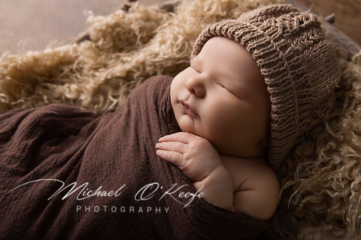 Sleeping newborn baby wrapped in brown cloth wearing a knit bonnet hat, professional newborn photography by Michael O'Keefe.