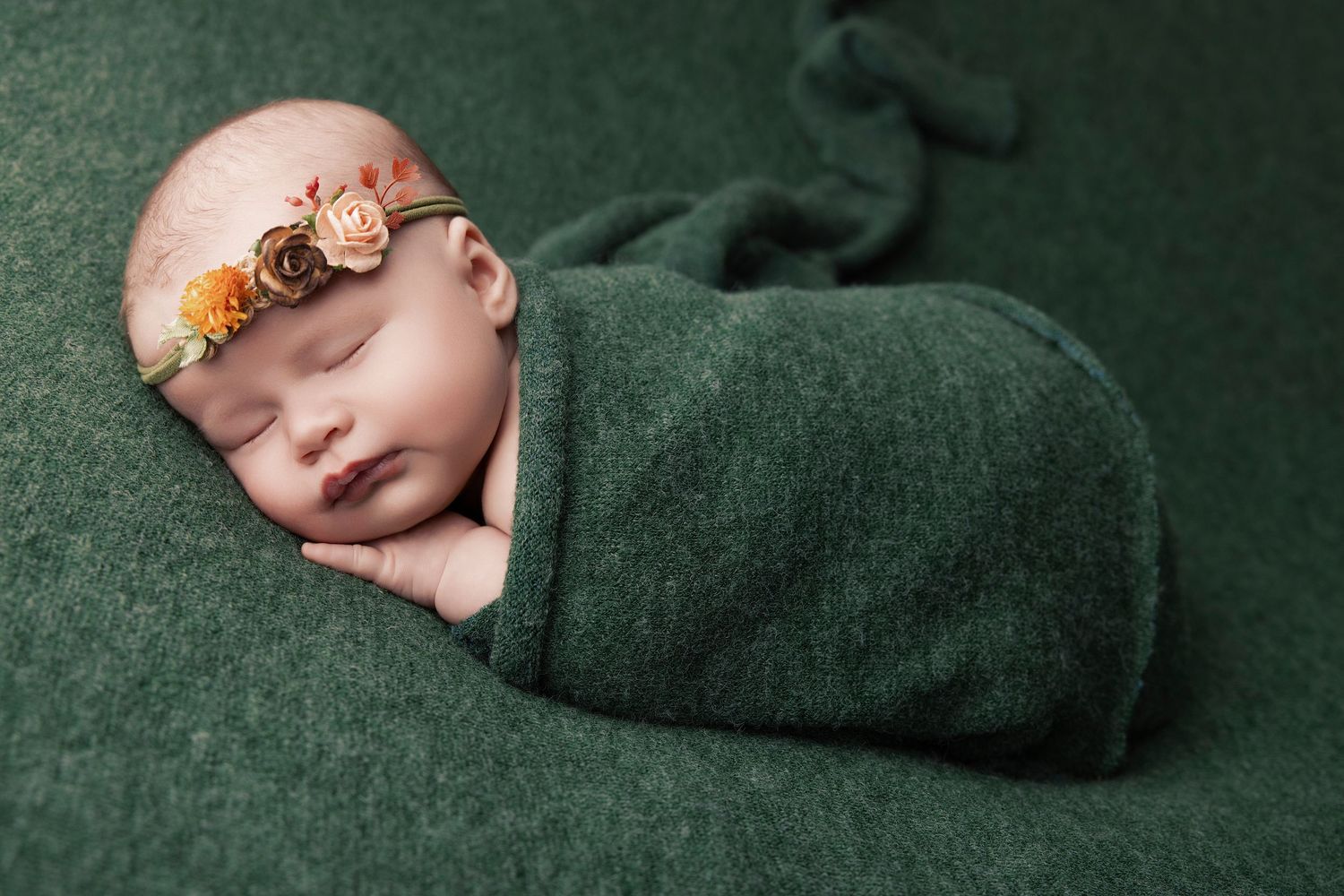 Sleeping newborn wrapped in green blanket wearing floral headband on dark green backdrop.