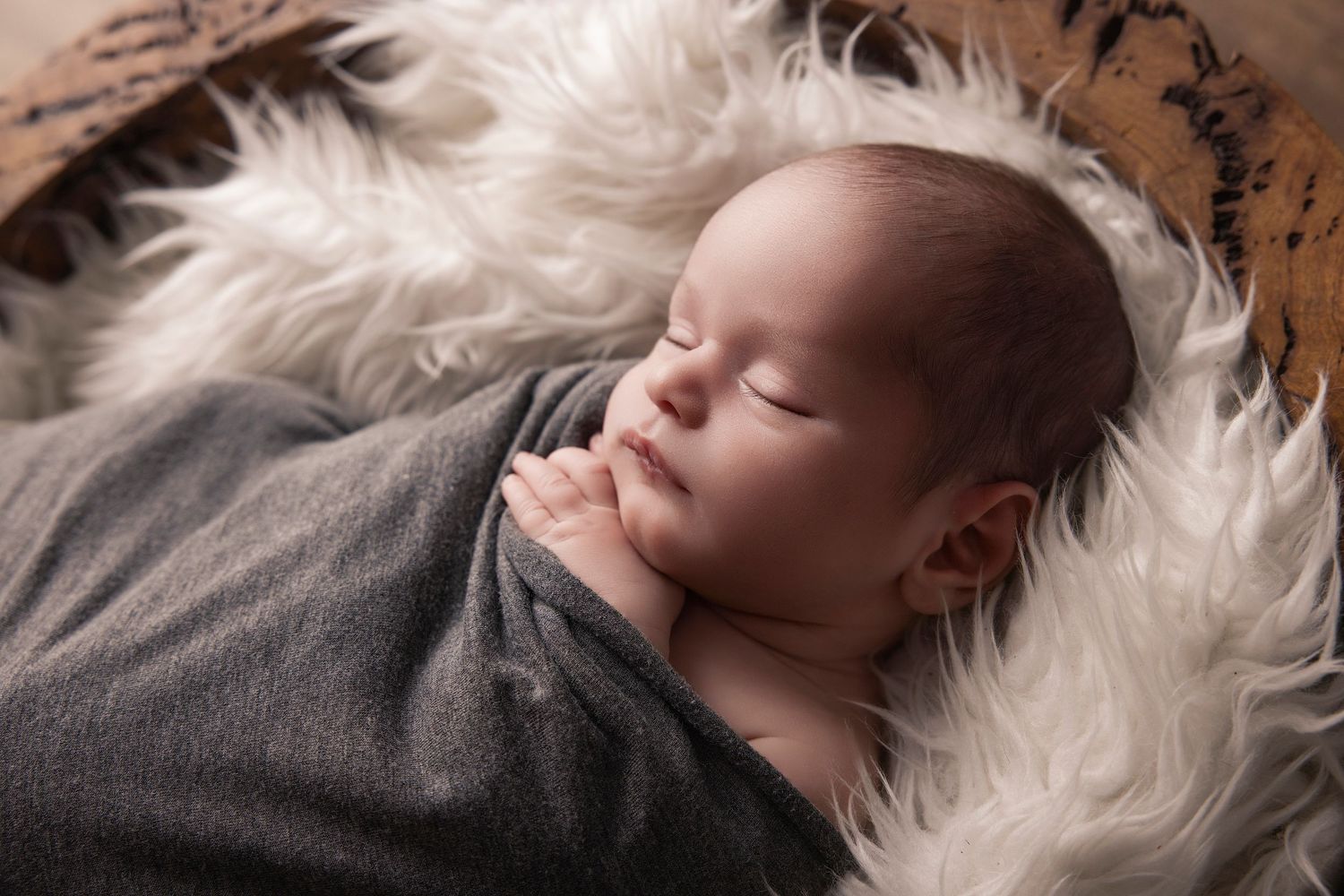 Peaceful sleeping baby wrapped in gray blanket resting on soft white fur backdrop during professional photo session.