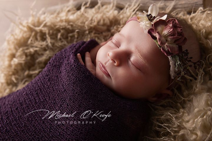 A newborn baby sleeping peacefully on soft beige fur blanket wearing a deep purple wrap in professional studio setting.