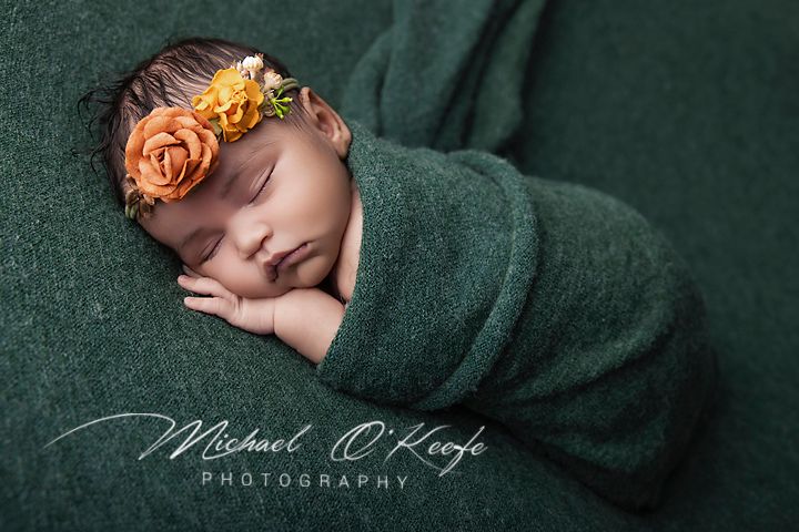 Newborn baby swaddled in green wrap with orange floral headband, sleeping peacefully in professional photography portrait.
