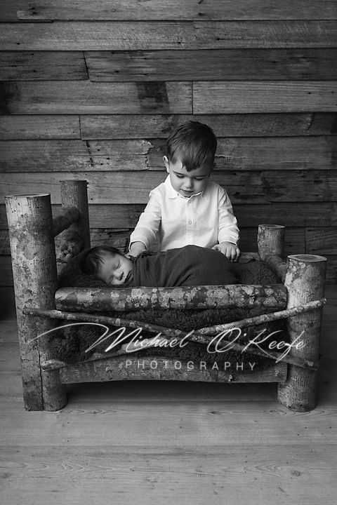 Black and white photo of a toddler boy watching over his swaddled newborn sibling on a rustic log bed.