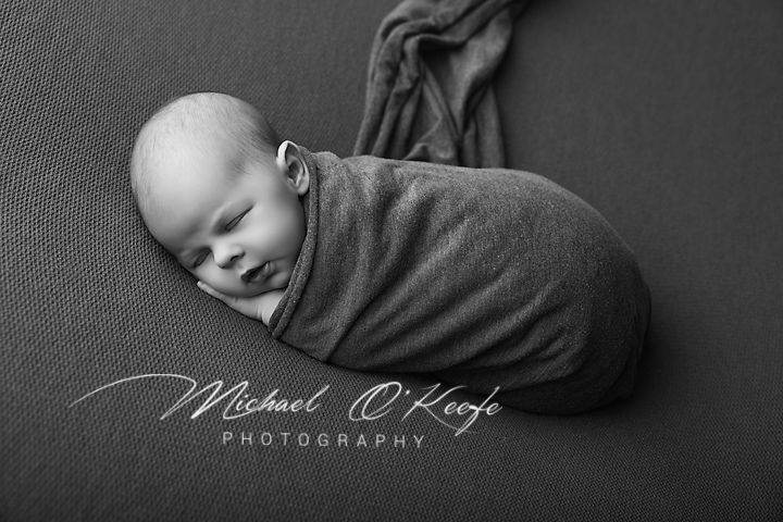 Black and white newborn photo of a sleeping baby swaddled in a gray wrap by Michael O'Keefe Photography.