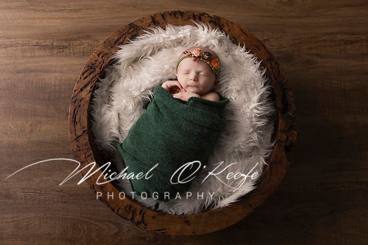 A newborn wrapped in green swaddle rests peacefully in a round wooden bowl lined with white fur.