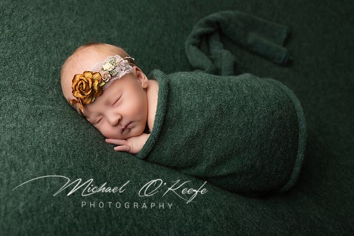 Newborn baby swaddled in green wrap with gold floral headband, sleeping peacefully on green backdrop.