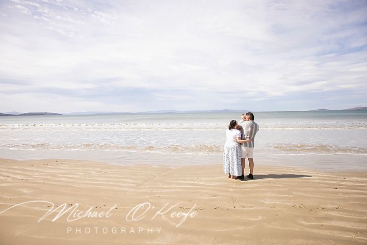 Couple sitting together on a sandy beach with rolling waves and cloudy sky in the background.
