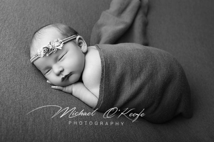 Black and white newborn photo of sleeping baby swaddled in soft wrap with floral headband by Michael O'Keefe Photography.