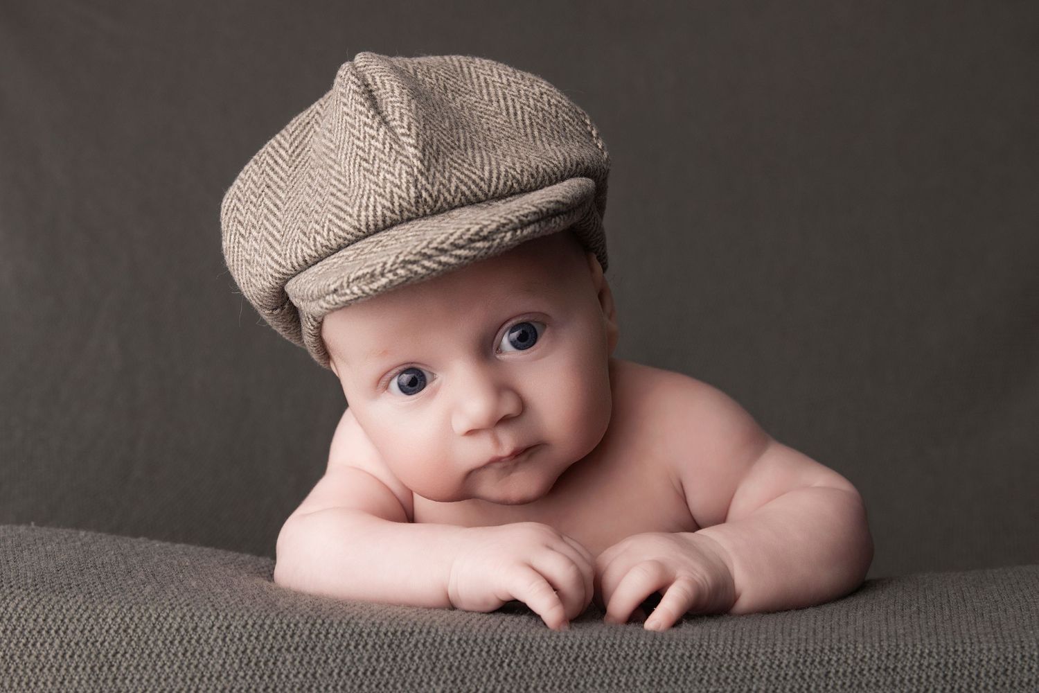 An artistic portrait series shows a baby in a newsboy cap laying on a gray backdrop with different poses.