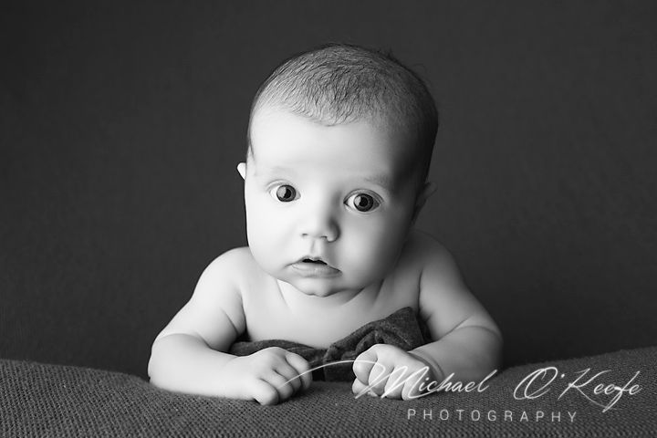 Black and white portrait of a curious baby lying on stomach, looking at camera by Michael O'Keefe Photography.