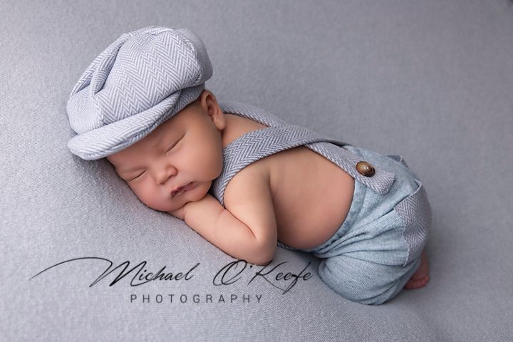 Newborn baby wearing soft blue outfit and gray cap sleeping peacefully on gray backdrop in professional photo shoot.