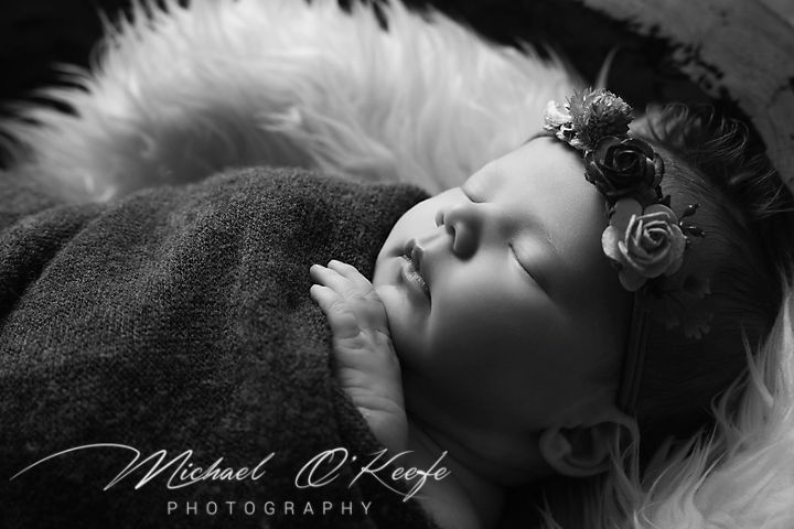 A black and white newborn portrait features a sleeping baby wearing a floral headband on soft fur bedding.