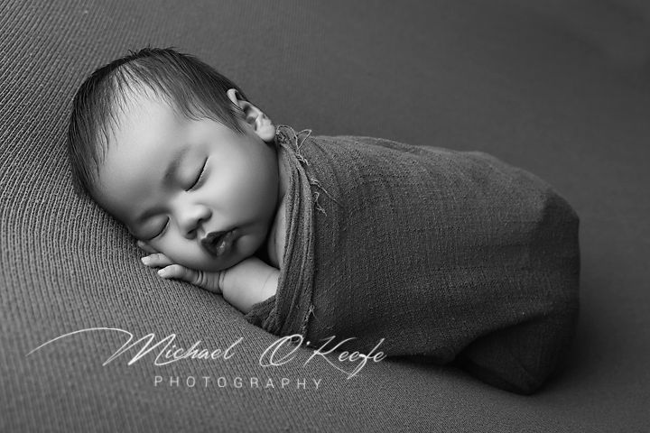 Black and white photo of a sleeping newborn baby swaddled in a soft wrap by Michael O'Keefe Photography.