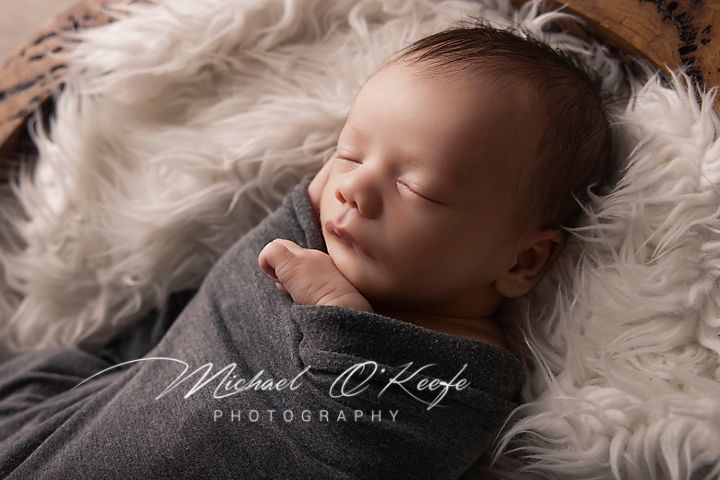 Peaceful newborn baby sleeping wrapped in gray swaddle on fluffy white fur, professional photography by Michael O'Keefe.