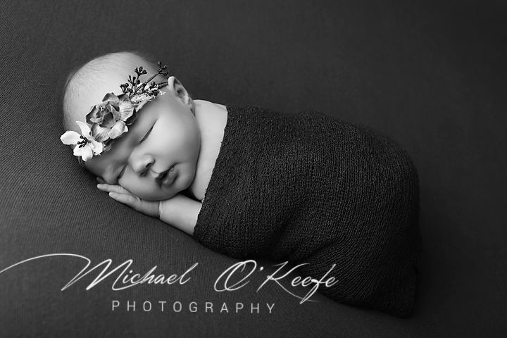Black and white newborn photography of a sleeping baby wrapped in dark knit fabric wearing a floral headband.