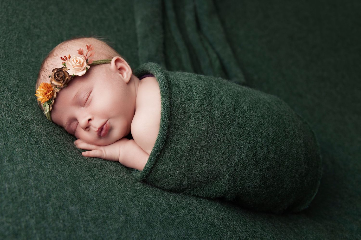 Sleeping newborn baby wrapped in soft dark green swaddle blanket during professional photography session.