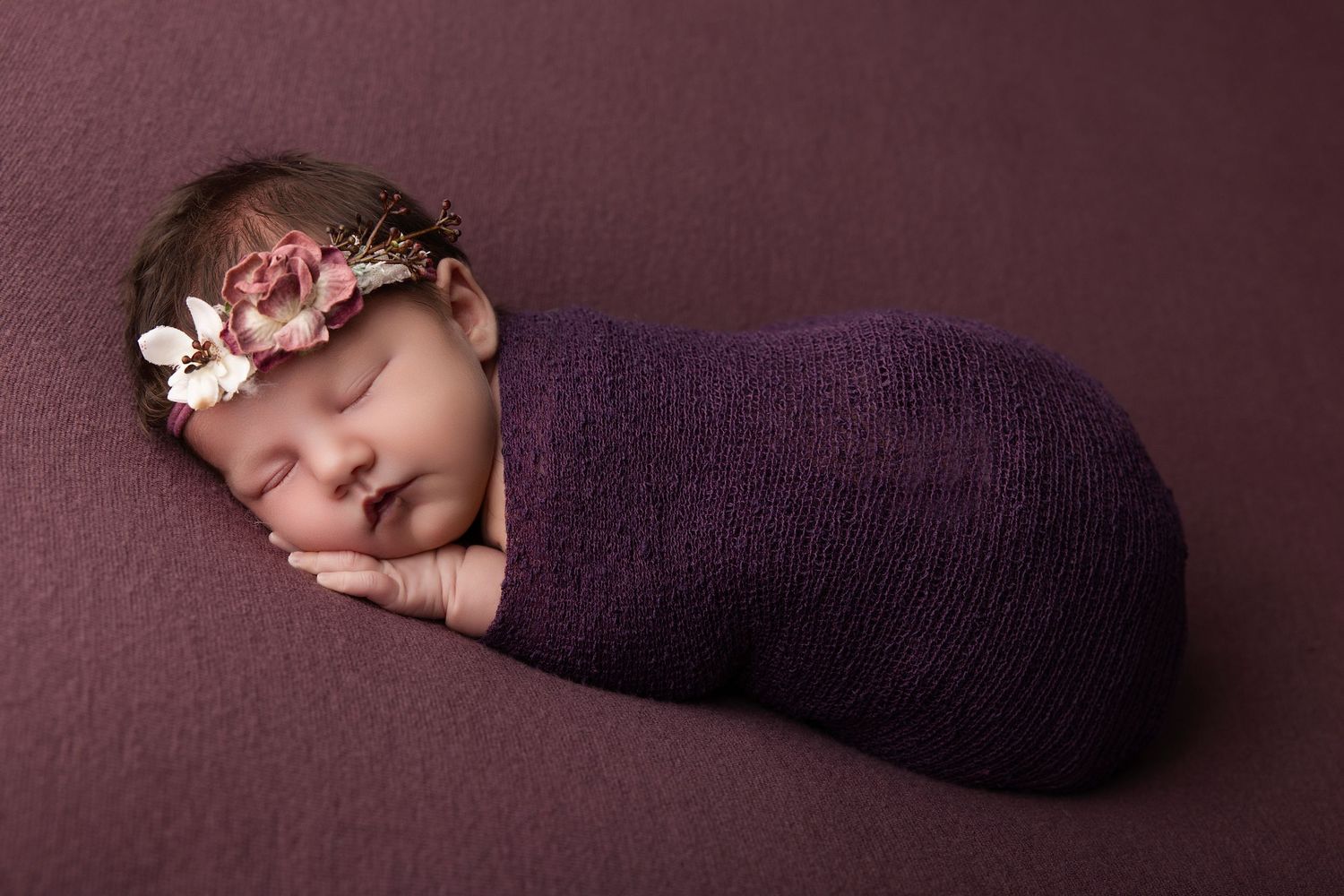 Newborn baby girl swaddled in purple wrap with floral headband, sleeping peacefully on matching purple backdrop.