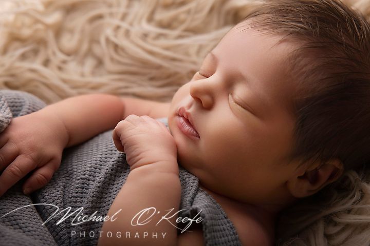 Peaceful newborn baby sleeping on a fluffy beige blanket, wrapped in a soft gray knit, by Michael O'Keefe Photography.