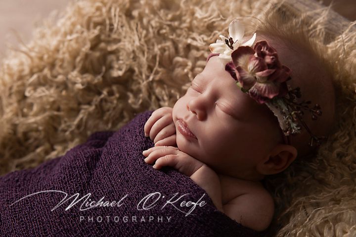 A newborn baby sleeps peacefully on soft beige fur while wrapped in a delicate knitted blanket.