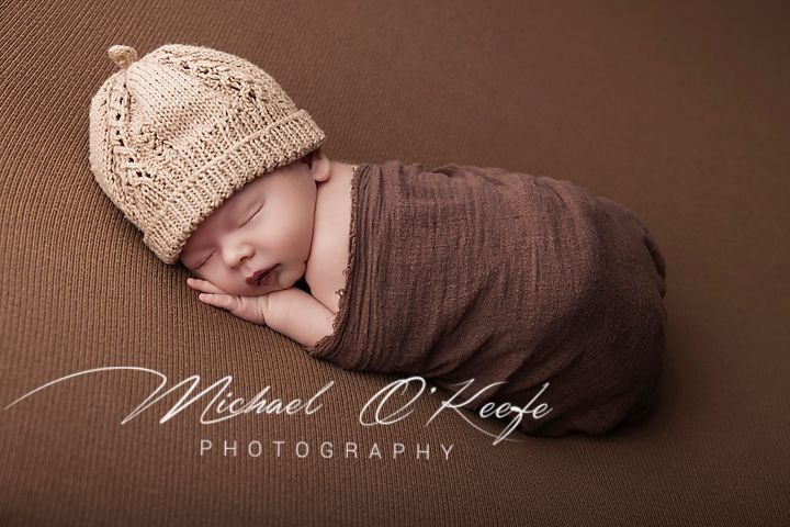 Newborn baby swaddled in brown wrap wearing a knit beige hat, sleeping peacefully on a brown backdrop.