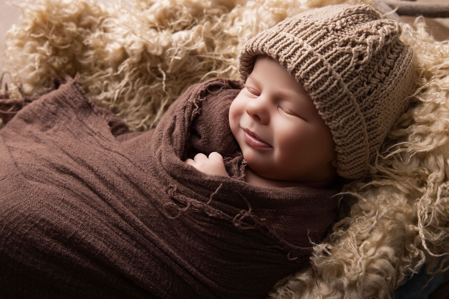 Smiling newborn baby wrapped in brown swaddle wearing knit beanie hat, sleeping peacefully on fluffy cream fur blanket.