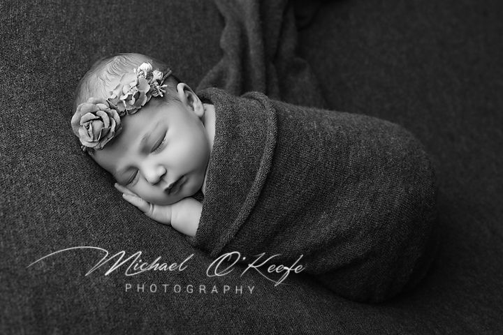 Black and white newborn photography of sleeping baby swaddled in gray wrap wearing floral headband by Michael OKeefe.