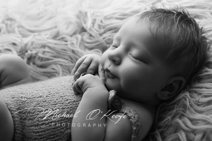 Black and white newborn photography of a sleeping baby with hands near face on a fluffy blanket by Michael O'Keefe.
