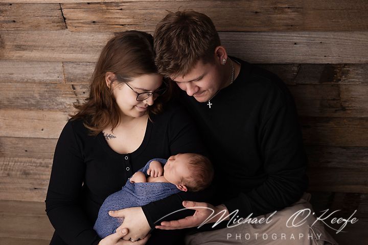 Family portrait of couple holding sleeping baby against rustic wooden wall.