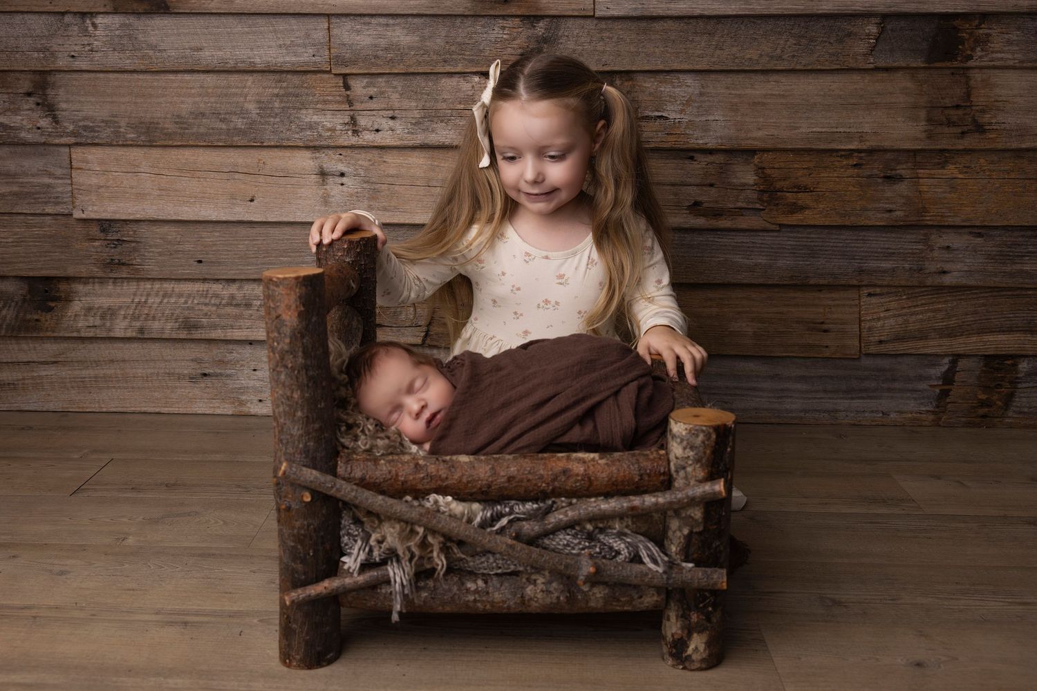 Rustic wooden baby bed with sleeping newborn and sibling in brown and cream tones.