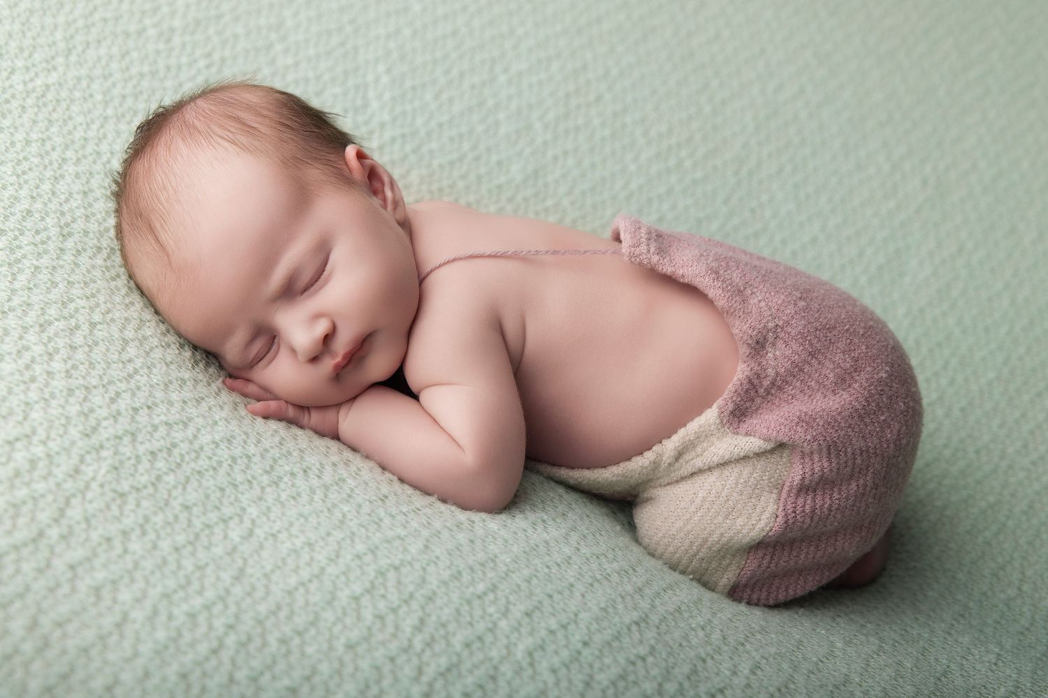 Sleeping newborn baby in pink knit shorts posed on a mint green blanket, smiling peacefully.