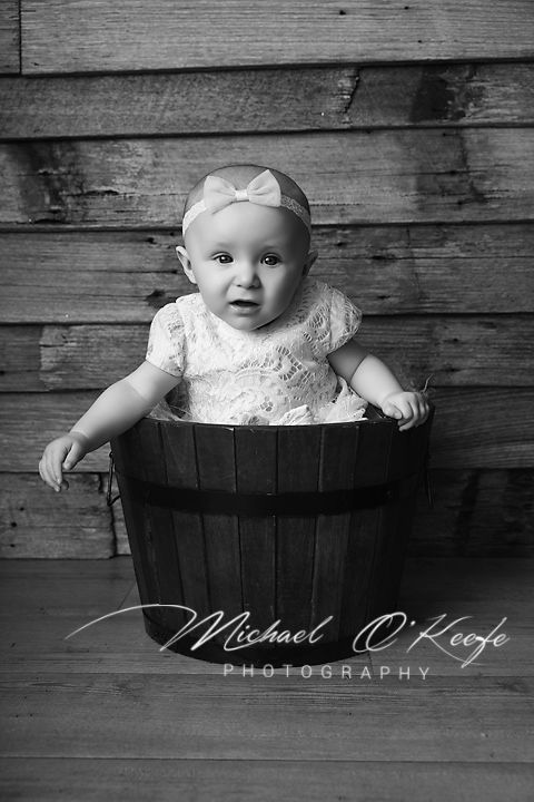 Black and white photo of a baby girl in a bow headband sitting in a wooden barrel bucket, by Michael O'Keefe Photography.