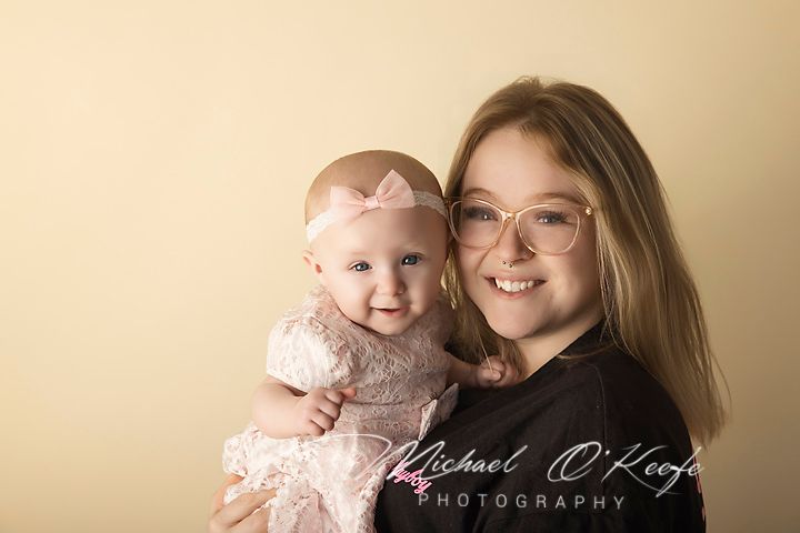 Older girl with glasses holding smiling baby in pink lace dress and bow headband, professional studio portrait.
