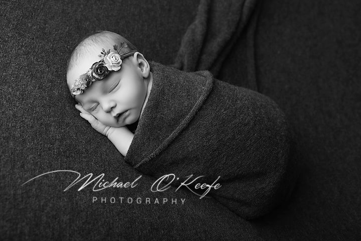 Black and white newborn photo of sleeping baby swaddled in dark wrap with floral headband by Michael O'Keefe Photography.