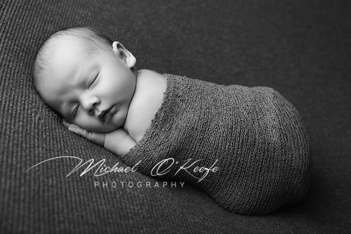 A sleeping newborn baby wrapped snugly in a gray knit blanket lies on a dark textured surface in black and white photography.