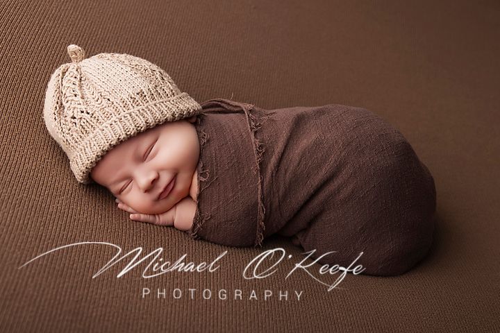 Smiling newborn baby wrapped in brown swaddle wearing knit beige hat, professional newborn photography by Michael O'Keefe.