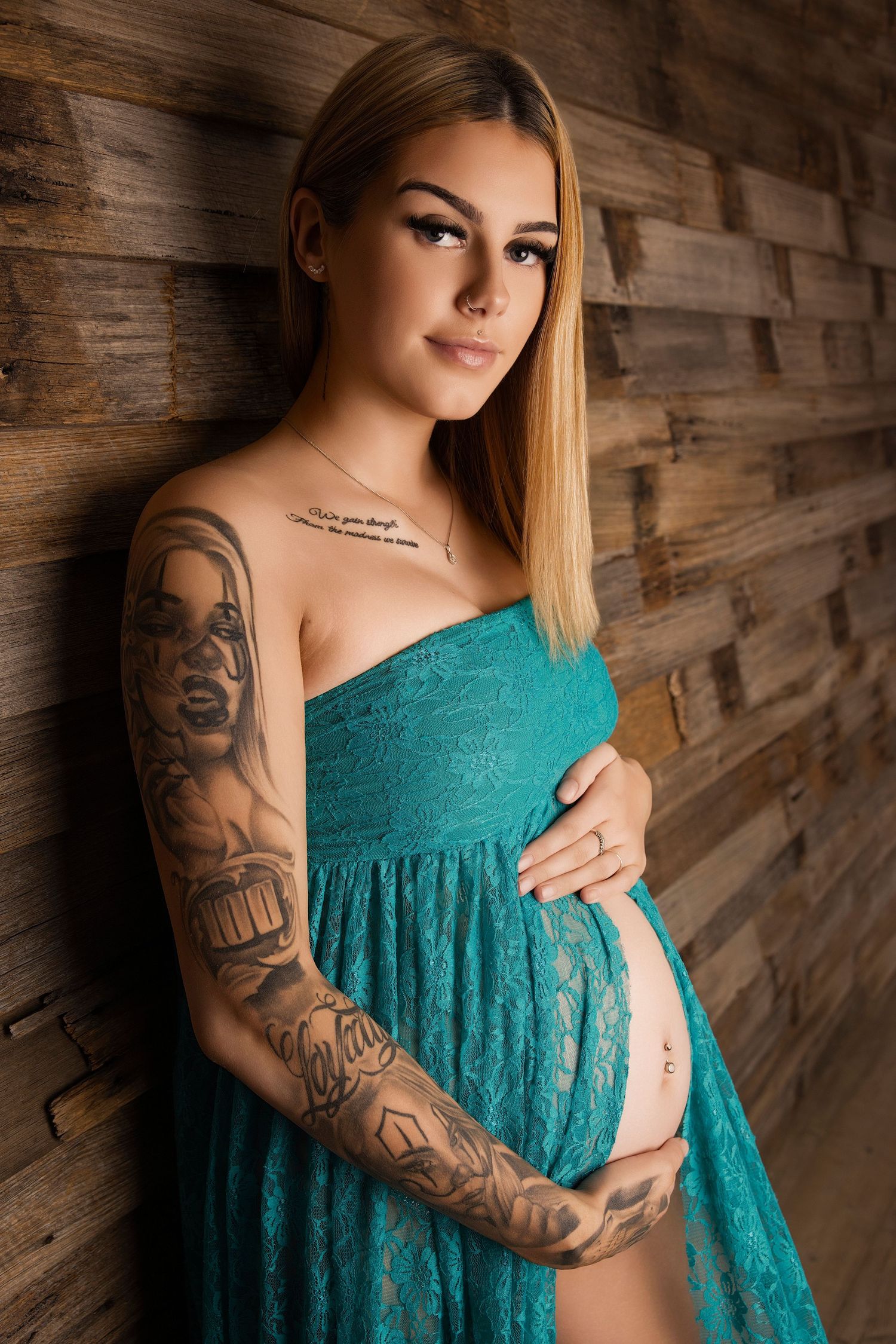 A maternity photoshoot against a rustic wood wall features a person in a turquoise lace dress with visible arm tattoos.