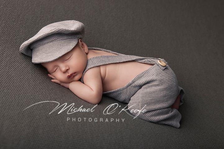 Sleeping newborn baby wearing a gray newsboy cap and suspender outfit posed on a gray backdrop.