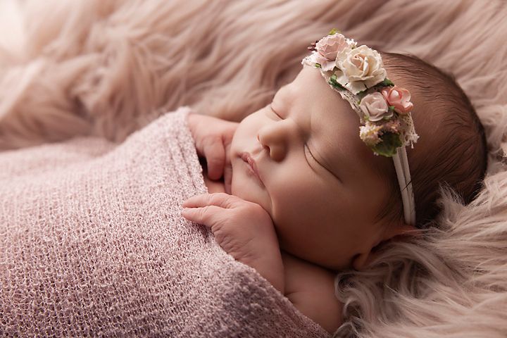 Sleeping baby wrapped in pink swaddle with delicate floral crown on fuzzy pink backdrop.