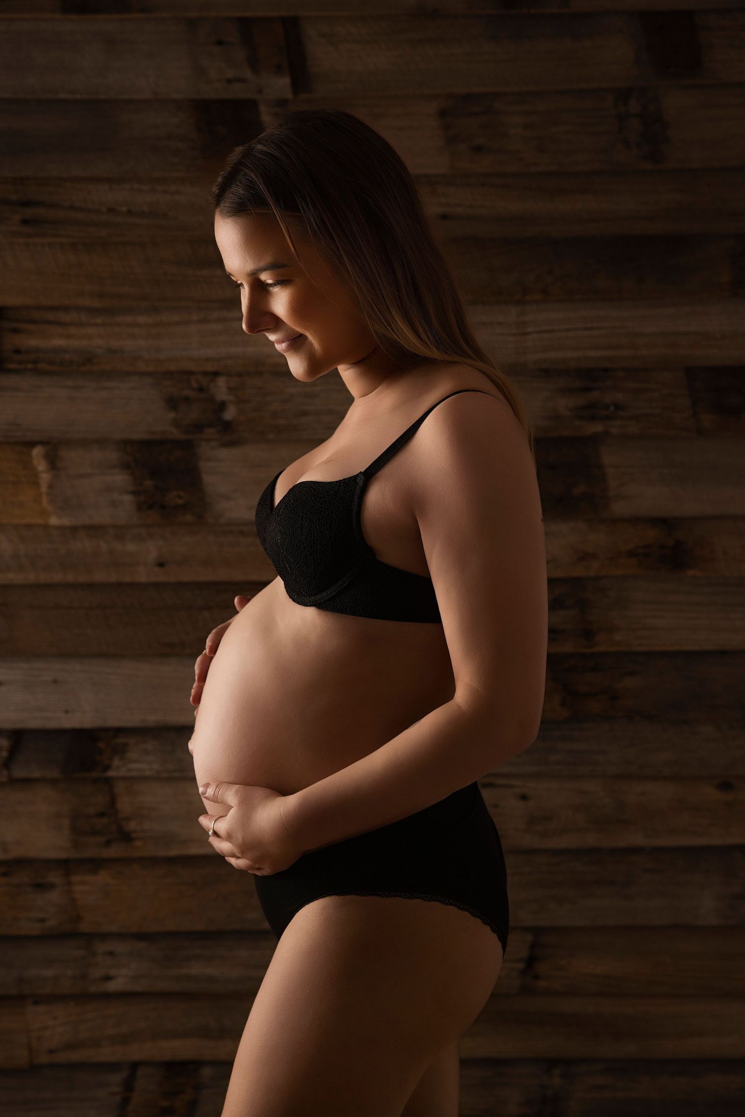 Maternity portrait of woman in black against rustic wooden wall with dramatic lighting.