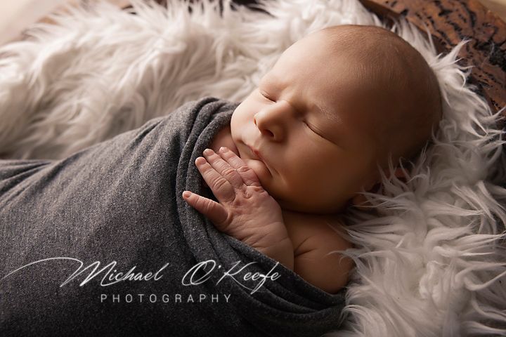 Newborn baby swaddled in gray wrap sleeping peacefully on fluffy white fur, photographed by Michael O'Keefe Photography.
