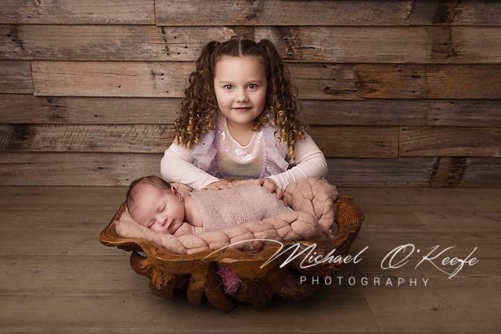 Young girl with curly hair smiles holding sleeping newborn in wooden bowl, professional newborn photography by Michael O'Keefe.
