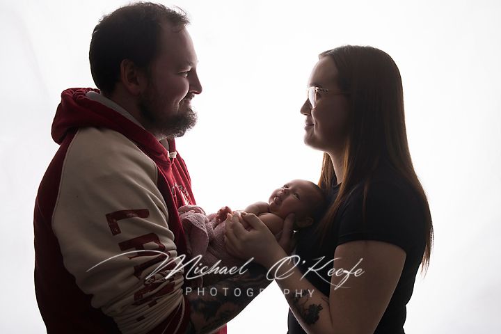 A couple gazes lovingly at their newborn baby in a softly lit studio portrait by Michael O'Keefe Photography.