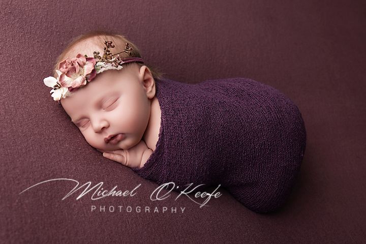 Newborn baby girl swaddled in purple wrap with floral headband, sleeping peacefully on purple backdrop.