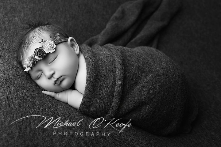 Black and white newborn portrait of a sleeping baby in a floral headband, wrapped in a textured blanket, by Michael O'Keefe Photography.