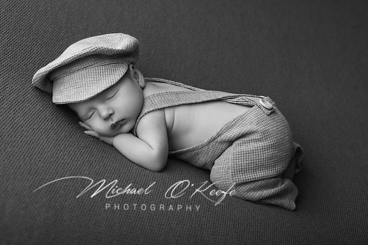 Sleeping newborn baby wearing a newsboy cap and suspenders outfit in black and white photography.