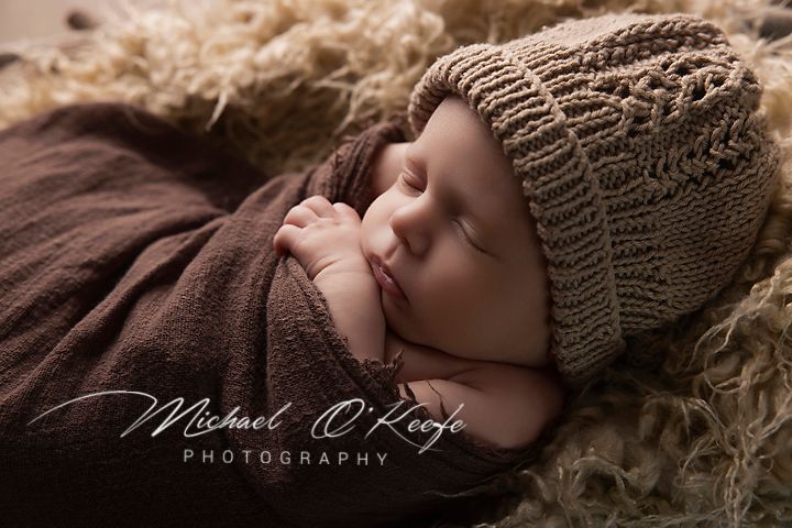 Newborn baby sleeping peacefully wrapped in brown knit blanket with matching hat by Michael O'Keefe Photography.