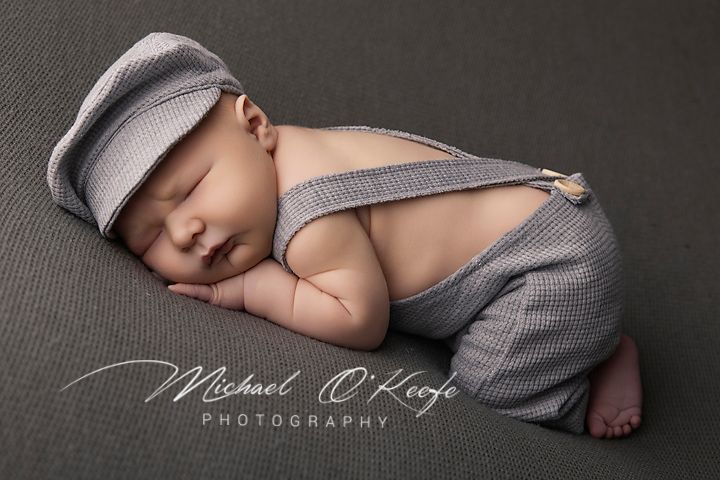 Sleeping newborn baby in gray suspender romper and newsboy cap posed on gray backdrop by Michael O'Keefe Photography.