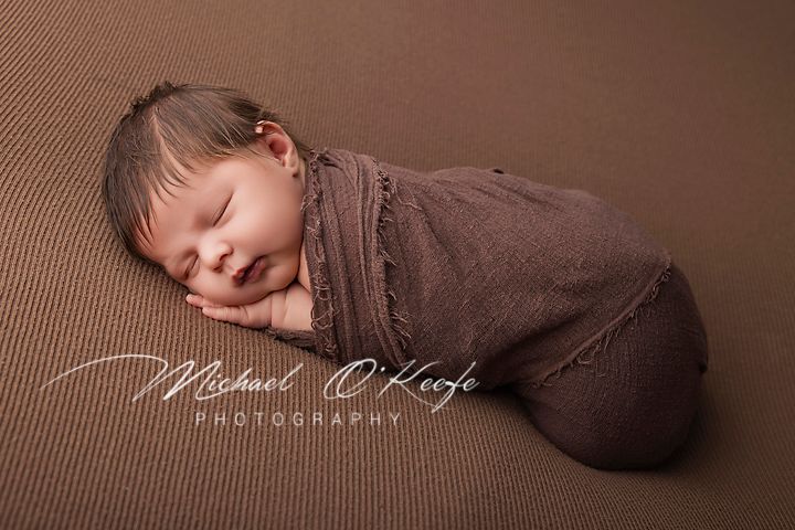A sleeping newborn baby wrapped in brown fabric on a textured brown backdrop in professional studio lighting.