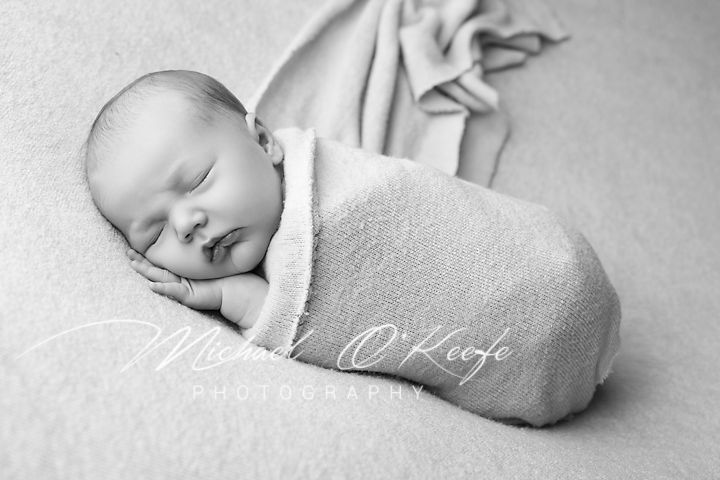 A sleeping newborn baby wrapped in a soft white blanket lies peacefully in black and white photography.